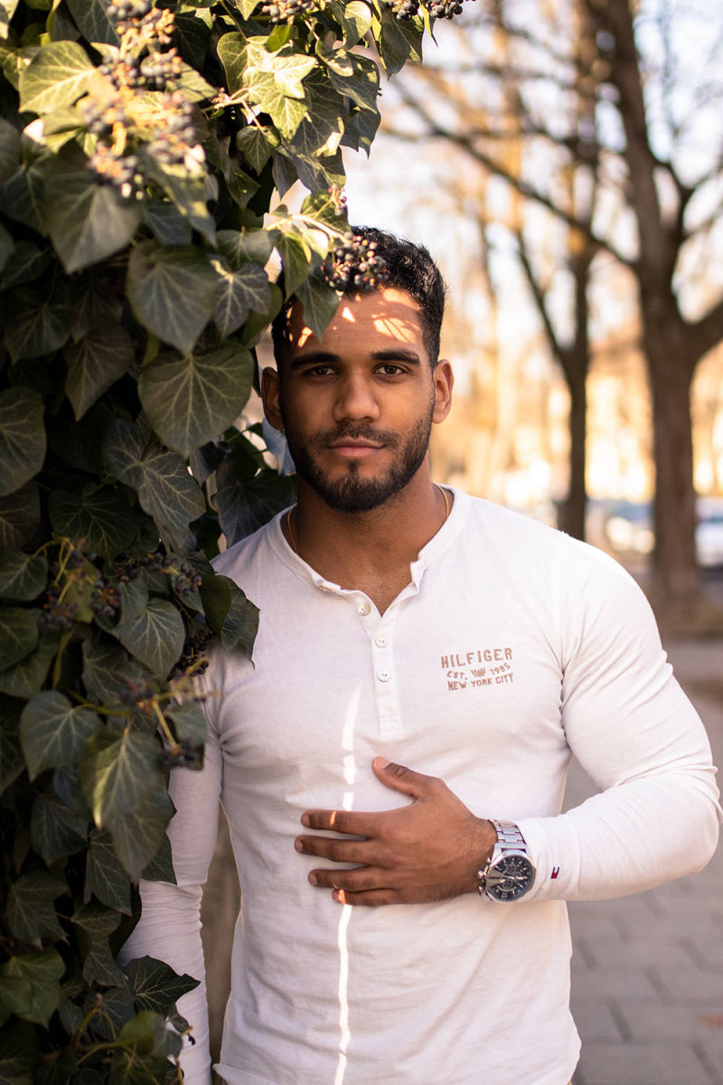 Junger Mann mit Bart wird fotografiert für Portraitfotos an einer Straße, er trägt ein weißes Hemd und eine Uhr und auf ihn fällt Sonnenlicht durch die Blätter der wachsenden Pflanzen