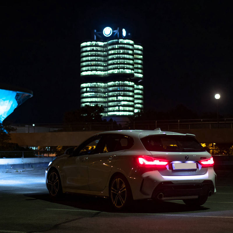 Ein 135er 1er BMW M Paket steht im Dunkeln mit leuchtenden Autoscheinwerfern auf dem Parkplatz gegenüber BMW-Welt und BMW-Vierzylinder Museum